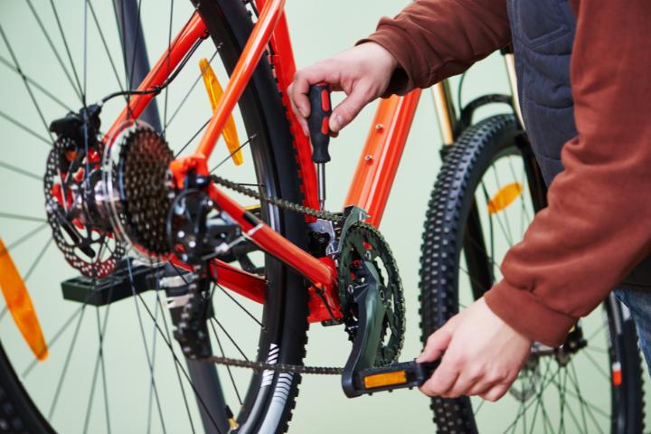 Close up of a person&#039;s hands and gears on a bicycling. One hand is holding a screwdriver into a bike chain, the other is holding the pedal of the bike.