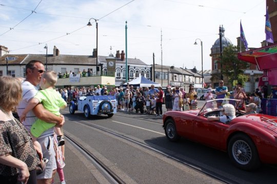 Parade of vintage cars driving down Fleetwood high street while onlookers watch from the sides.